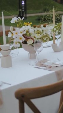 Dışarıdaki masada çiçekli vazo şenlikli bir akşam yemeği için. Düğün dekoru, cam, çiçekler, mumlar. Güzel bir etkinlik için dekorasyon. Tahta sandalyeler, düğün yemeği. Dikey video. Yüksek kalite 4k