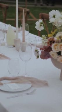 Dikey video. Parti ya da düğün için dekore edilmiş bir masa. Açık hava ziyafeti, çiçekli masa, mumlar, sandalyeler, küçük rahat bir düğün partisinde beyaz tabaklar, yiyecek yok. Ağır çekim. Yüksek kalite 4k görüntü