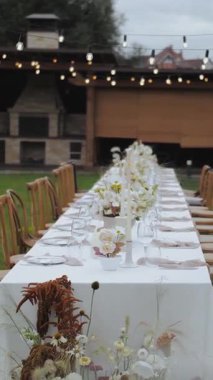 Dikey video. Bir düğünde ya da başka bir etkinlikte, bir vazo ve açık havada bir buket taze çiçekle süslenmiş bir yemek masasının önünde masayı aydınlatan bir çelenk. Yüksek