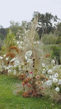 Dikey video. Çiçek aranjmanı, çiçek dekoru, çimlerin üzerindeki çiçekler. Boho tarzı düğün kemeri. Kurumuş çiçekler ve yeşil çimlerde pastel çiçekler. Parkta düğün töreni.