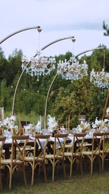 Dikey video. Kristal avizelerle süslenmiş düğün masası, mumlar ve kurutulmuş çiçekler Boho düğün yemeği için, tabaklar ve şarap bardakları, insan yok. Sokak dekoru. Yüksek kalite 4k görüntü