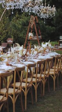 Dikey video. Mumlar, tahta sandalyeler, çiçekler, tabaklar ve açık havada kristal avizelerle süslenmiş bir masa. Boho düğünü, insan yok. Düğün partisi. Yüksek kalite 4k görüntü