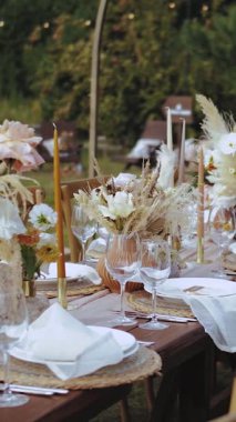 Masa dekorasyonu, mumlar, kurumuş çiçekler ve pastel çiçeklerle dekorasyon. Boho tarzı açık hava düğünü, tabaklar ve bardaklar, insan ya da yemek yok, ağır çekim. Dikey video. Yüksek kalite 4k görüntü