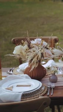 Dikey video. Açık hava şenlikli bir akşam yemeği için çok güzel dekore edilmiş bir masa. Boho düğünü, beyaz tabaklar, bardaklar, insan yok ve ahşap bir masada yemek. Yüksek kalite 4k görüntü