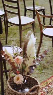 Dikey video. Yeşil çimlerin üzerinde açık hava düğün dekoru. Pastel çiçek aranjmanları, kurutulmuş çiçekler, tahta sandalyeler ve halı. Ağır çekim. Yüksek kalite 4k görüntü