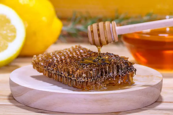 Close-up of a dispenser dripping honey on a piece of honeycomb placed on a wooden plate, with a bowl of honey, a sprig of rosemary, a lemon and a half and a real honeycomb in the background