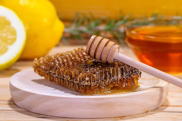 Close-up of a piece of honeycomb and a dispenser on a wooden plate, with a bowl of honey, a rosemary sprig, a lemon and a half and a real honeycomb in the background