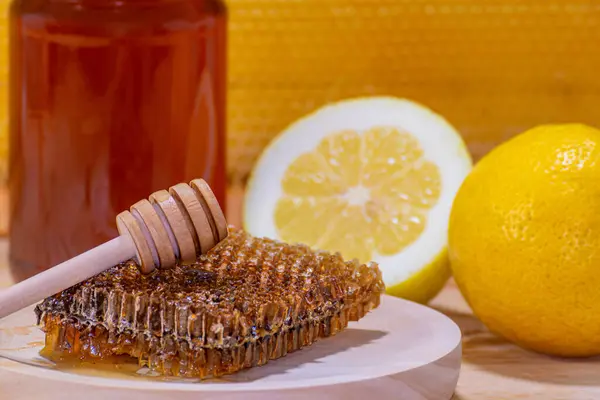 Close-up of a piece of honeycomb and a dispenser on a wooden plate, with a jar of honey, a lemon and a half and a real honeycomb in the background
