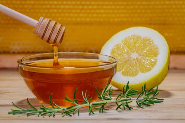 Dispenser dripping honey on a bowl, a rosemary sprig and half a lemon on wood with a real honeycomb in the background