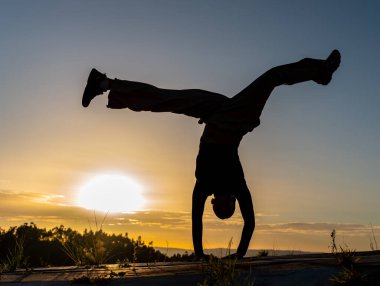 Gün batımında yüz üstü break dans yapan bir kadının arka ışıklandırılmış silueti. Günbatımı, gökyüzü bir zemin gibi, sıcak bir sahne ekler.
