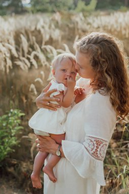 Bir aile doğanın ortasında iki çocuk, ikiz kız kardeş, kollarında kızı olan bir anne ve açık renk giysiler içinde yürür.