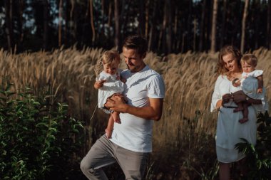 Bir aile doğanın ortasında iki çocuk, ikiz kız kardeş ile yürür; ebeveynler çocuğu ellerinde tutar ve uzun otların ortasında yürür.