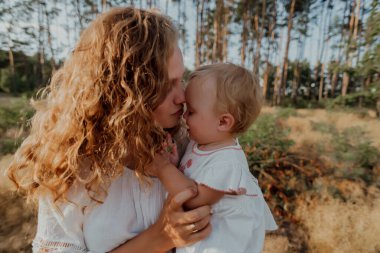 Bir aile doğanın ortasında iki çocuk, ikiz kız kardeş, kollarında kızı olan bir anne ve açık renk giysiler içinde yürür.