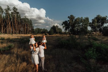 Bir aile doğanın ortasında iki çocuk, ikiz kız kardeş ile yürür; ebeveynler küçük çocuklarla oynar ve onları ellerinin başının üstünde tutarlar; aile hafif giysiler giyer.