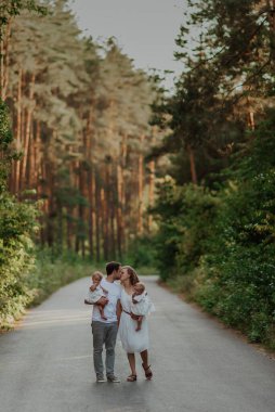 Bir aile doğanın ortasında iki çocuk, ikiz kız kardeş ile yürür; oğlanlar ormanın ortasında asfaltlanmış yol boyunca yürür kollarında çocuklarıyla; aile hafif kıyafetler giyer; baba anneyi öper; 