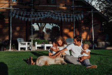 Bir aile doğanın ortasında iki çocuk, ikiz kız kardeş ile yürüyor; ebeveynler evlerinin yakınında yeşil bir çimenlikte çocuklarıyla oynuyorlar ve köpekleri onların yanında.
