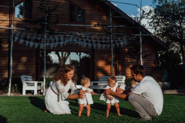 Bir aile doğanın ortasında iki çocuk, ikiz kız kardeş ile yürüyor; ebeveynler evlerinin yakınında yeşil bir çimenlikte çocuklarıyla oynuyorlar ve köpekleri onların yanında.