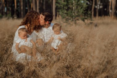 Bir aile doğanın ortasında iki çocukla yürür, ikiz kız kardeşler; ebeveynler çim yapraklarının arkasında çocuklarıyla öpüşürler; ailenin arka planında keskin çimler vardır.