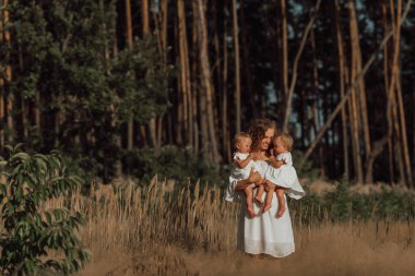 a family walks in the middle of nature with two children, twin sisters; mother holds two little twin daughters in her arms;  dressed in light clothes clipart