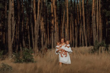 Bir aile doğanın ortasında iki çocuk, ikiz kız kardeş ile yürür; anne iki küçük ikiz kızını kucağında tutar; açık renk kıyafetler giyer.