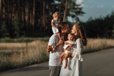 Bir aile doğanın ortasında iki çocuk, ikiz kız kardeş ile yürür; ebeveynler çocukları kollarında tutar; baba anneyi öper