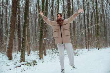 Karlı bir ormanda bir kız karda sevinir; kapüşonlu bir ceket ve bir eşarp ve spor ayakkabı giyiyor