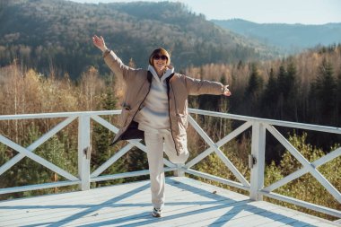 Sıcak elbiseli bir kız dağların arka planında terasta durur; güneşli havada zıplar ve neşelenir.