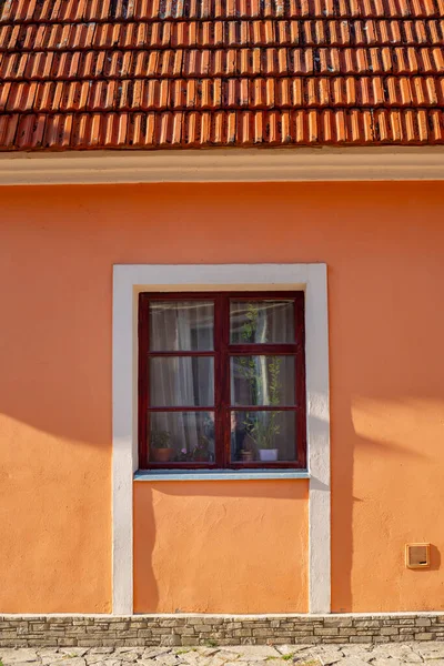 Part of the decoration of the facade of the building and the material from which it is built;  a window with flowerpots and a wall of an orange-colored house with a roof made of tiles