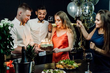 Şenlik masasındaki arkadaşlar doğum günü kızını kutluyorlar; alkol ve atıştırmalıklar masada; kırmızı şarap ellerinde; mutlu ve neşeli gülümsemeler olan bardaklar; kırmızı elbiseli bir kız pastadaki mumları üflüyor