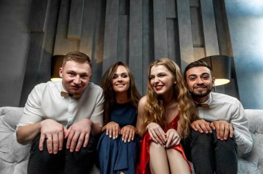 Stüdyoda bir kanepede şarap kadehleriyle oturuyorlar; gri bir koltukta poz veriyorlar; birbirlerine yakın oturuyorlar ve birbirlerine gülümsüyorlar ve kameraya suratlarını ekşitiyorlar.