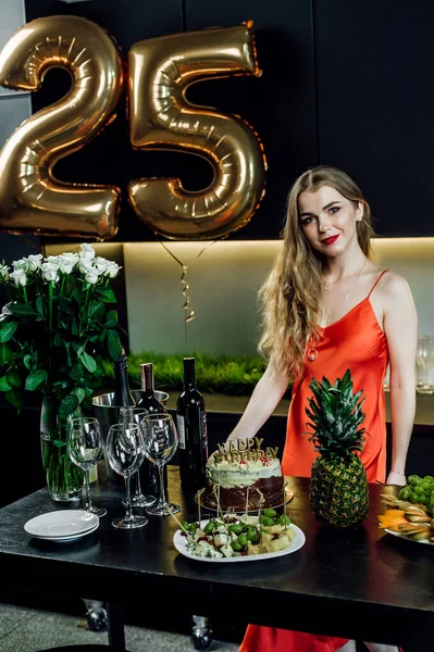 A beautiful girl in a red dress is standing near the festive table;Friends at the festive table congratulate the birthday girl;alcohol and snacks are on the table;glasses with red wine in hands;