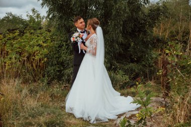 Yeni evliler düğün günlerinde bir fotoğrafçıya doğanın arka planına karşı poz verirler; adam koyu klasik bir ceket giyer ve kadın beyaz bir elbise giyer; çok mutlu ve neşeli bir çift aşık olur.