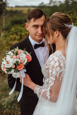 Yeni evliler düğün günlerinde bir fotoğrafçıya doğanın arka planına karşı poz verirler; adam koyu klasik bir ceket giyer ve kadın beyaz bir elbise giyer; çok mutlu ve neşeli bir çift aşık olur.
