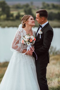 Yeni evliler düğün günlerinde bir fotoğrafçıya doğanın arka planına karşı poz verirler; adam koyu klasik bir ceket giyer ve kadın beyaz bir elbise giyer; çok mutlu ve neşeli bir çift aşık olur.