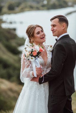 Yeni evliler düğün günlerinde bir fotoğrafçıya doğanın arka planına karşı poz verirler; adam koyu klasik bir ceket giyer ve kadın beyaz bir elbise giyer; çok mutlu ve neşeli bir çift aşık olur.