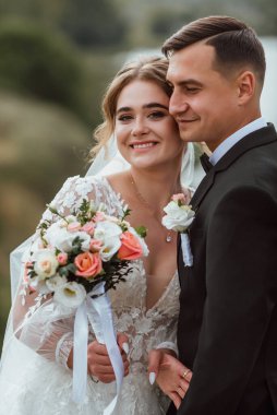 Yeni evliler düğün günlerinde bir fotoğrafçıya doğanın arka planına karşı poz verirler; adam koyu klasik bir ceket giyer ve kadın beyaz bir elbise giyer; çok mutlu ve neşeli bir çift aşık olur.