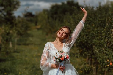 Genç ve güzel bir gelin, doğanın arka planına karşı bir düğün buketi ile poz verir; kız beyaz bir gelinlik giyer; düğün gününde bir an; mutlu kız duygularını ifade eder