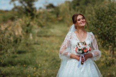 Genç ve güzel bir gelin, doğanın arka planına karşı bir düğün buketi ile poz verir; kız beyaz bir gelinlik giyer; düğün gününde bir an; mutlu kız duygularını ifade eder