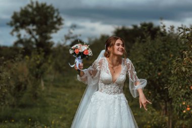 Genç ve güzel bir gelin, doğanın arka planına karşı bir düğün buketi ile poz verir; kız beyaz bir gelinlik giyer; düğün gününde bir an; mutlu kız duygularını ifade eder