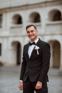 Genç ve yakışıklı bir damat mimarinin arka planına karşı poz verir; adam klasik siyah bir düğün kıyafeti giyer; düğün gününde an; mutlu damat duygularını ifade eder