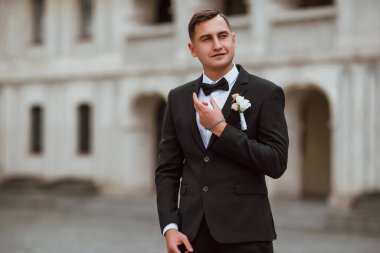 Genç ve yakışıklı bir damat mimarinin arka planına karşı poz verir; adam klasik siyah bir düğün kıyafeti giyer; düğün gününde an; mutlu damat duygularını ifade eder