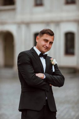 Genç ve yakışıklı bir damat mimarinin arka planına karşı poz verir; adam klasik siyah bir düğün kıyafeti giyer; düğün gününde an; mutlu damat duygularını ifade eder