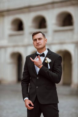 Genç ve yakışıklı bir damat mimarinin arka planına karşı poz verir; adam klasik siyah bir düğün kıyafeti giyer; düğün gününde an; mutlu damat duygularını ifade eder