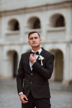 Genç ve yakışıklı bir damat mimarinin arka planına karşı poz verir; adam klasik siyah bir düğün kıyafeti giyer; düğün gününde an; mutlu damat duygularını ifade eder
