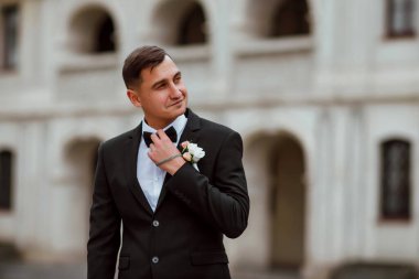 Genç ve yakışıklı bir damat mimarinin arka planına karşı poz verir; adam klasik siyah bir düğün kıyafeti giyer; düğün gününde an; mutlu damat duygularını ifade eder