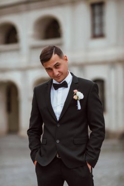 Genç ve yakışıklı bir damat mimarinin arka planına karşı poz verir; adam klasik siyah bir düğün kıyafeti giyer; düğün gününde an; mutlu damat duygularını ifade eder