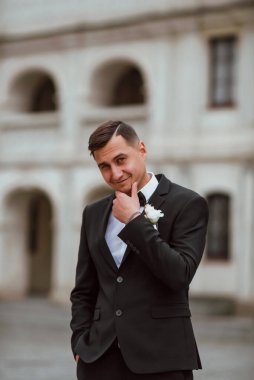 Genç ve yakışıklı bir damat mimarinin arka planına karşı poz verir; adam klasik siyah bir düğün kıyafeti giyer; düğün gününde an; mutlu damat duygularını ifade eder