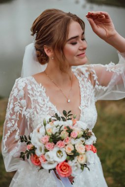 Genç ve güzel bir gelin, doğanın arka planına karşı bir düğün buketi ile poz verir; kız beyaz bir gelinlik giyer; düğün gününde bir an; mutlu kız duygularını ifade eder