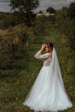 Genç ve güzel bir gelin, doğanın arka planına karşı bir düğün buketi ile poz verir; kız beyaz bir gelinlik giyer; düğün gününde bir an; mutlu kız duygularını ifade eder