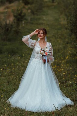 Genç ve güzel bir gelin, doğanın arka planına karşı bir düğün buketi ile poz verir; kız beyaz bir gelinlik giyer; düğün gününde bir an; mutlu kız duygularını ifade eder